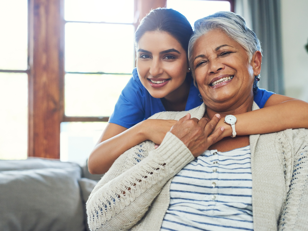 caregiver-giving-support-to-elderly A smiling elderly woman sits comfortably on a couch while a cheerful caregiver in blue scrubs stands behind her, wrapping her arms around the senior in a warm, supportive embrace. The bright, cozy living room in the background emphasizes the caring environment. This image represents compassionate senior home care in Bourne, highlighting the strong bond and trust between caregivers and seniors.