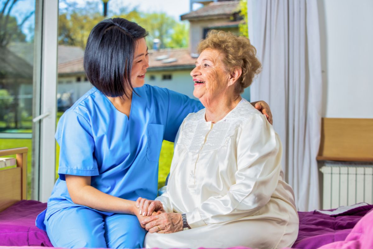 A caregiver laughs and holds the hand of an elderly woman setting on a bed.