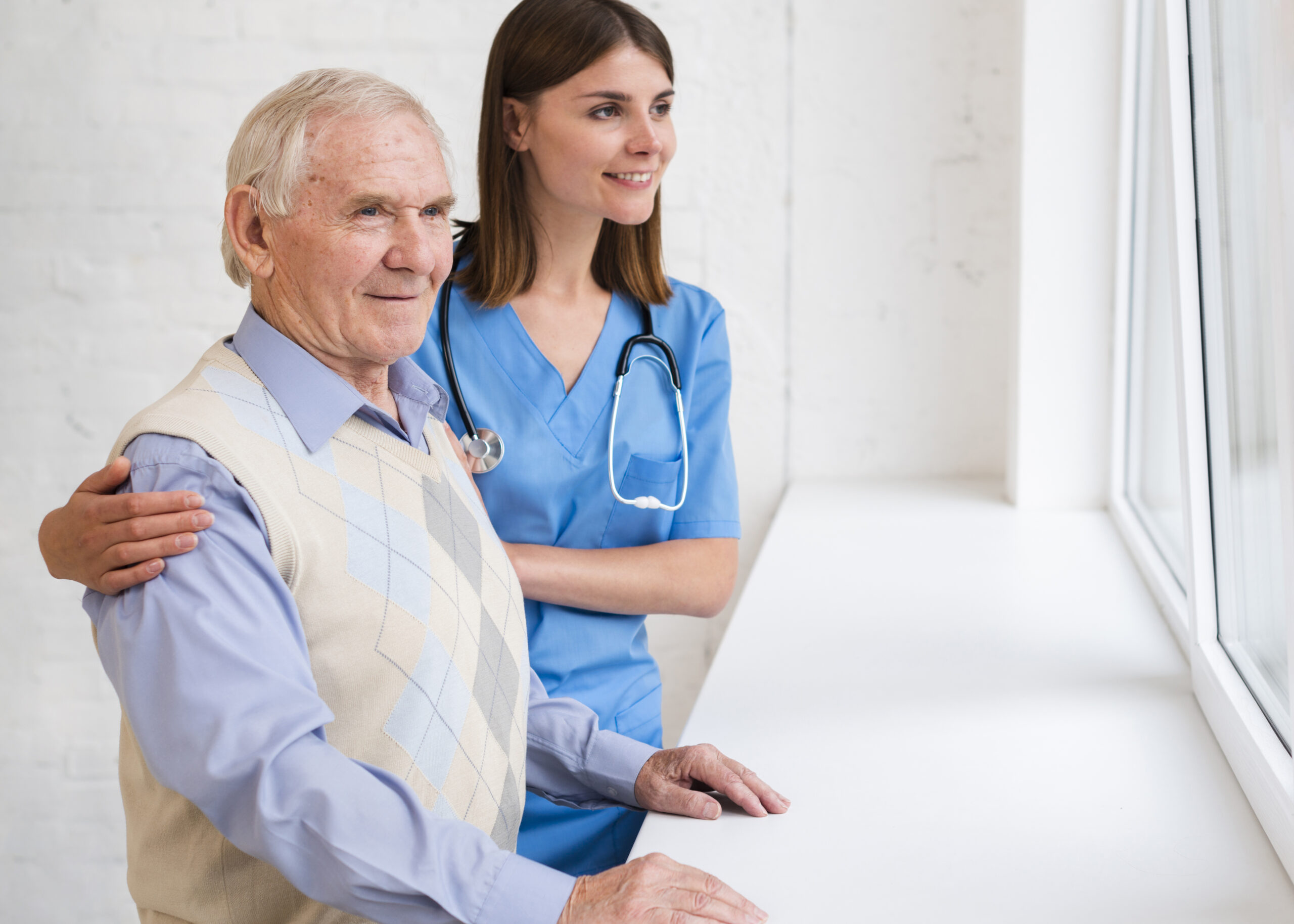 A caregiver in blue scrubs, wearing a stethoscope, stands beside an elderly person in a sleeveless sweater as they both look out of a window, sharing a quiet, reflective moment together.
