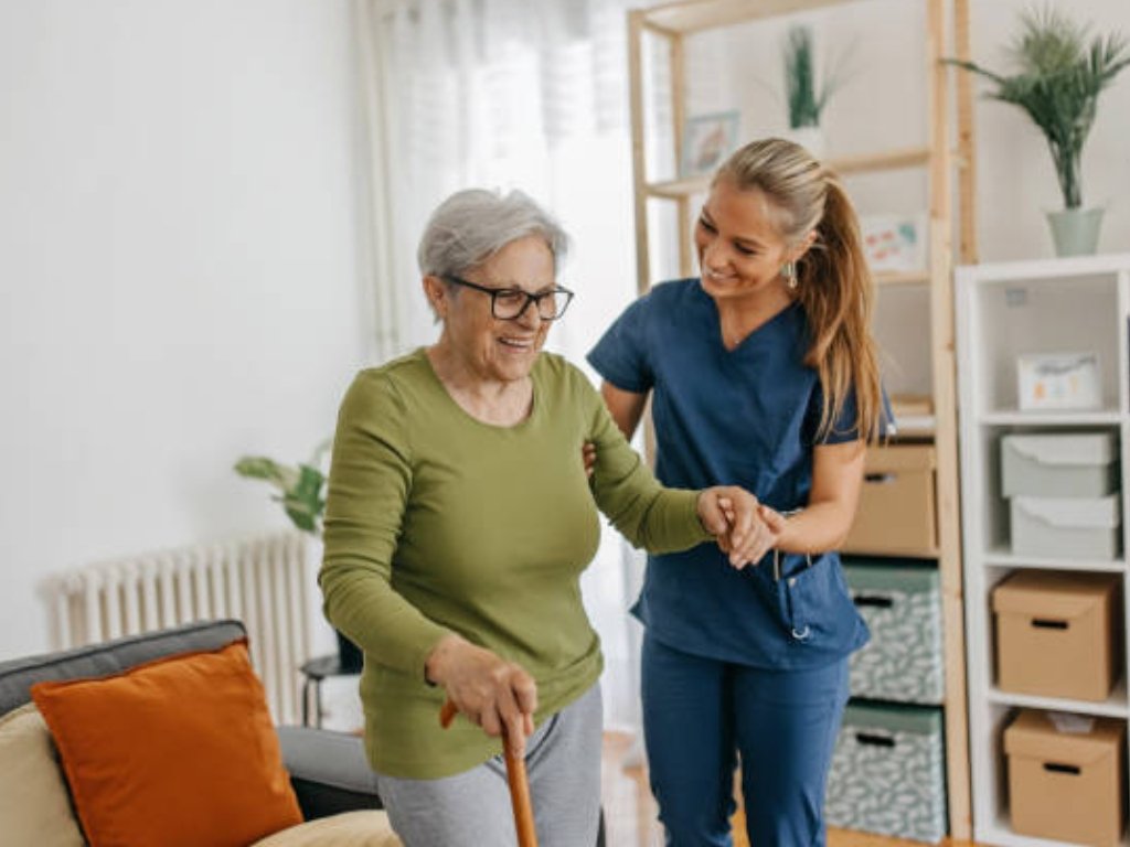 senior home care in New Bedford A smiling caregiver in blue scrubs assists an elderly woman with a cane inside a cozy, well-lit living room. The warm interaction reflects quality senior home care in New Bedford, emphasizing support, comfort, and independence for older adults.
